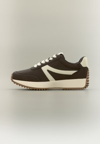Καφέ αθλητικό παπούτσι με μπεζ λεπτομέρειες, επάνω μέρος από υφασμάτινη υφή, στρογγυλεμένη μύτη, χοντρή, οδοντωτή σόλα και επίπεδα κορδόνια. Κομψός, μοντέρνος σχεδιασμός.