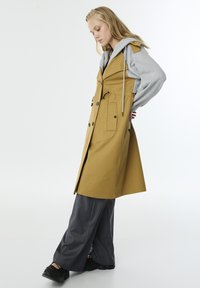 Langer senfgelber Trenchcoat mit Kapuze, doppelten Knöpfen und Seitentaschen, kombiniert mit grauen Jogginghosen und schwarzen Schuhen.