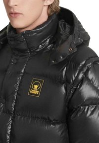 Giacca imbottita nera con finitura lucida, caratterizzata da un colletto alto, bottoni a pressione e una toppa logo gialla con un elefante sul petto.