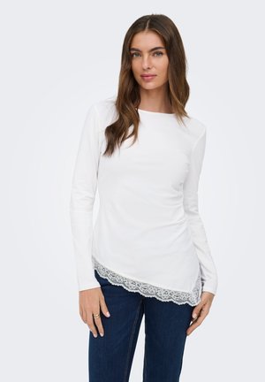 Femme aux longs cheveux bruns, portant un haut blanc à manches longues avec une bordure en dentelle à l'ourlet et un jean bleu foncé, debout devant un fond uni.