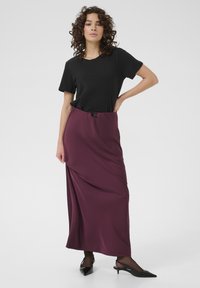 Schwarzes T-Shirt kombiniert mit einem knöchellangen burgunderroten Rock, der ein drapiertes Design und einen Tunnelzugbund aufweist. Schwarze spitze Absatzschuhe runden den Look ab.