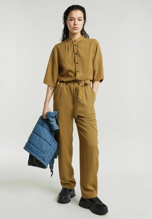 Frau trägt ein senfgelbes Hemd mit Knöpfen und Hose, hält eine blaue Steppjacke, steht mit einer Hand in der Tasche vor einfarbigem Hintergrund.