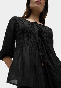 Schwarze Bluse mit geraffter Textur und weichem Stoff. Sie verfügt über einen Frontbindverschluss, lange Ärmel und eine fließende Silhouette.