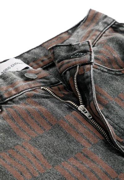 Jean en denim gris avec des rayures abstraites marron, fermeture éclair métallique partiellement ouverte, bouton et étiquette blanche visible.