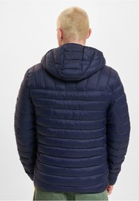 Mørkeblå pufferjakke med horisontalt quiltet mønster, elastisk hette og lett teksturert stoff. Har glidelås og tettsittende mansjetter.
