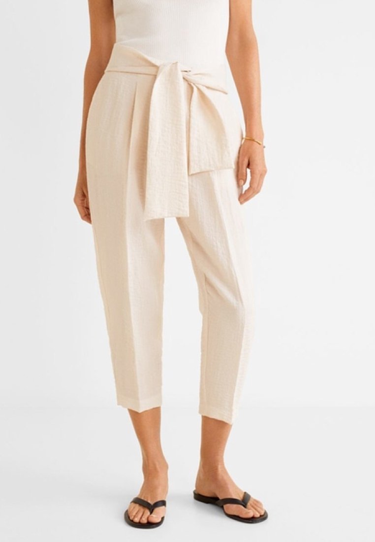 Femme portant un pantalon court taille haute beige clair avec une large ceinture en tissu nouée à l’avant et des sandales tongs noires.