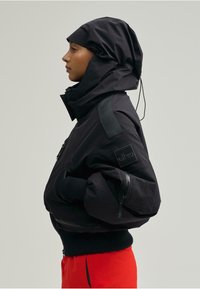 Fekete dzseki magas gallérral és kapucnival, cipzáras zsebekkel, bordázott mandzsettákkal és oldalsó logófolttal. Sima, matt textúrájú.