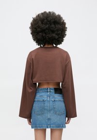 Rjav top z dolgimi rokavi, odrezan do pasu, kombiniran zmodro jeans mini krilo z raztrganimi robovi in črnim logotipnim našitkom na pasu.