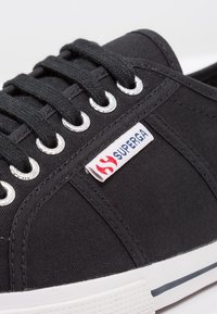Fekete vászon sportcipő ezüst fűzőlyukakkal, fekete fűzőkkel és fehér gumitalppal, piros és kék Superga logó címkével.