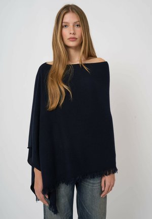 Młoda kobieta z długimi blond włosami, ubrana w luźne, ciemnoniebieskie ponczo z opuszczonymi ramionami oraz niebieskie dżinsy, stojąca na tle jednolitego, jasnego tła.