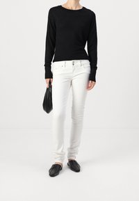 Pull noir avec des perles incorporées, jeans blancs slim-fit, et mocassins noirs plats, tenant un sac à main noir. Design simple et ajusté.