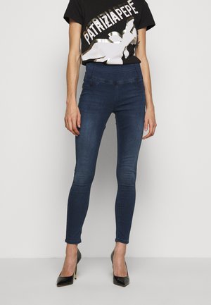 Kvinde iført mørkeblå højtaljede skinny jeans, sorte højhælede sko og en sort t-shirt med hvidt og gyldent grafisk tekst- og designmotiv.