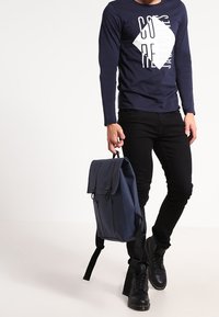 Námořnicky modrý batoh s překlopeným víkem, nastavovacími popruhy a patentkami; nošený osobou v námořnicky modrém triku s dlouhým rukávem a černých džínách.