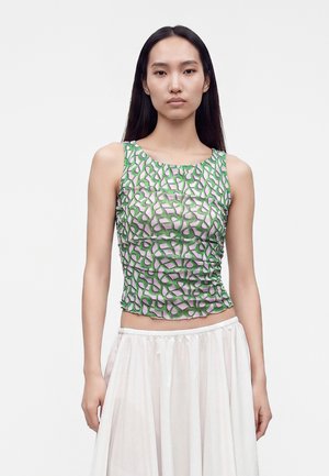 Jeune femme aux longs cheveux foncés portant un haut sans manches avec un motif abstrait vert et rose et une jupe blanche sur un fond uni.