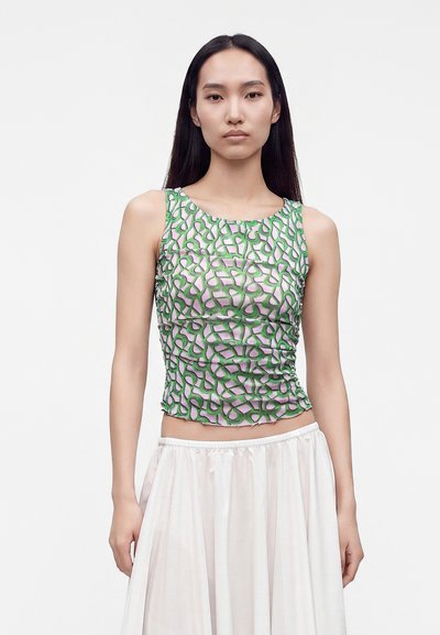 Jeune femme aux longs cheveux foncés portant un haut sans manches avec un motif abstrait vert et rose et une jupe blanche sur un fond uni.