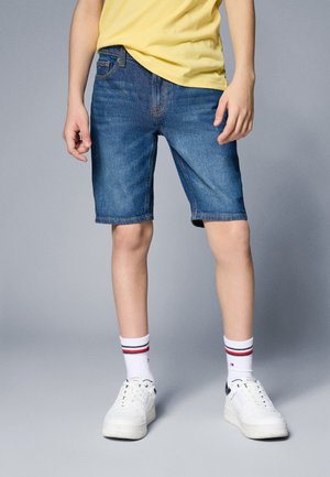 Personne portant un short en jean bleu, un t-shirt jaune, des baskets blanches et des chaussettes blanches avec des rayures rouges et bleu marine.