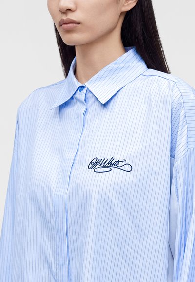 Femme portant une chemise bleu clair à fines rayures, avec col et logo brodé "Off White" sur la poitrine.