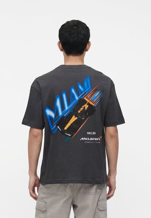 Osoba nosiaca čierne tričko s modrým textom "MIAMI" a grafikou McLaren Formula 1 auta na chrbte, stojaca pred jednoduchým pozadím.