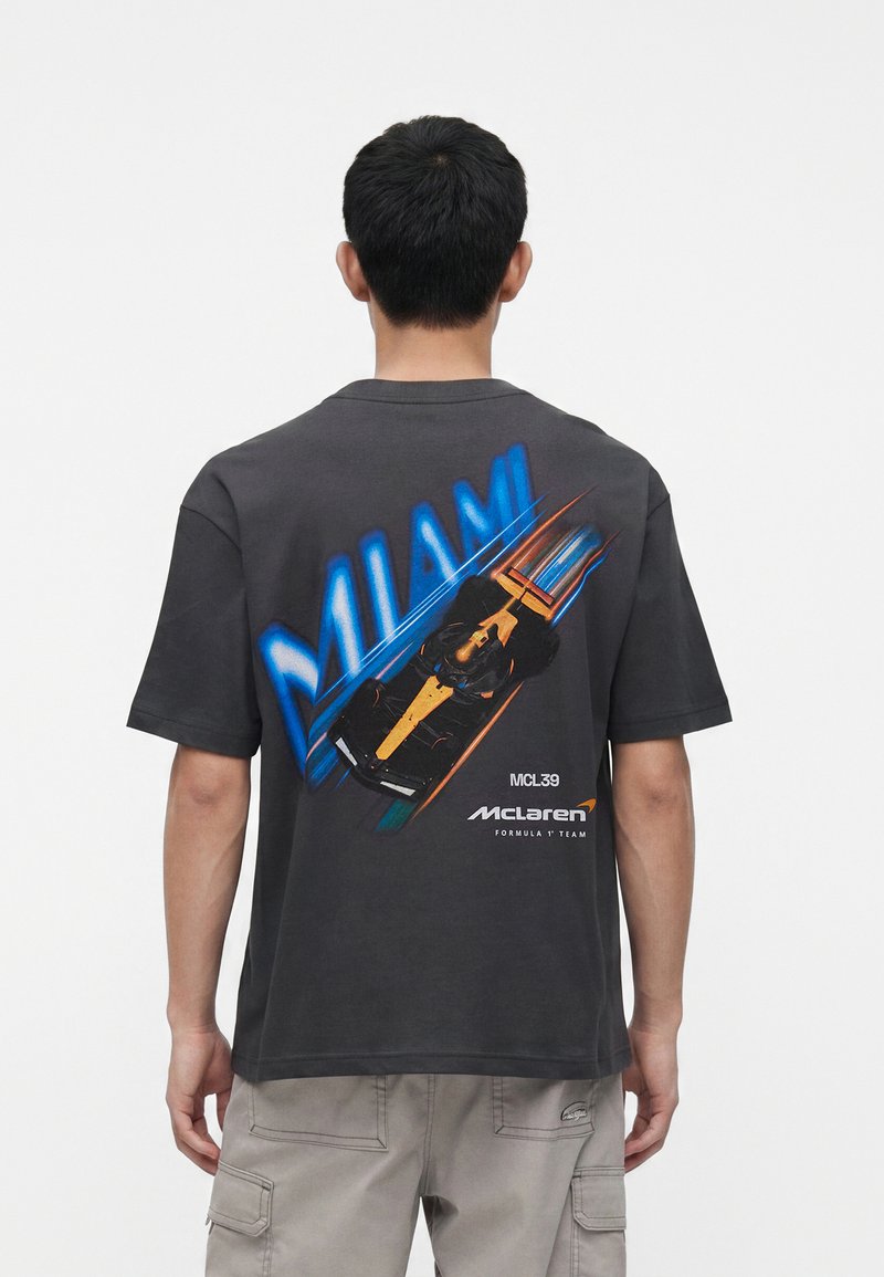 Άτομο που φοράει μαύρο T-shirt με μπλε κείμενο "MIAMI" και γραφικό αυτοκινήτου McLaren Formula 1 στην πλάτη, στέκεται μπροστά σε απλό φόντο.