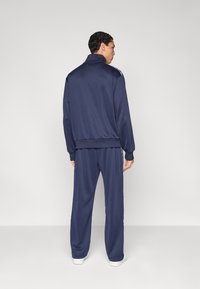 Μπλε σκούρο φόρμα από απαλό ύφασμα, με μπουφάν υψηλού γιακά και φαρδιά παντελόνια με ρίγες στο πλάι, σε συνδυασμό με λευκά παπούτσια.