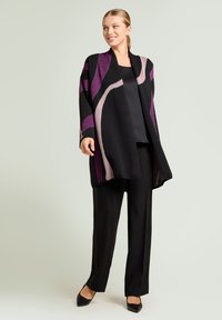 Zwarte cardigan met paarse en roze abstracte patronen, gecombineerd met een zwarte top en zwarte wijde broek met een ontspannen pasvorm en een open voorkant.