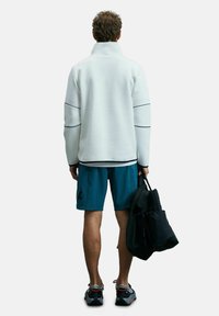 Veste en polaire blanche à col montant avec des coutures bleu marine, short turquoise, sac noir et chaussures de sport grises avec des accents rouges. Vue de dos.