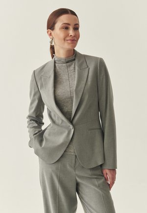 Kvinde med brunt hår i en lav hestehale iført sølvøreringe, en grå blazer, matchende bukser og en grå rullekravetrøje, stående med den ene hånd i lommen.