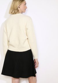 Ženska s kratkimi blond lasmi, oblečena v smetanast pulover in črno krilo, obrača se bočno proti enobarvni beli podlagi.
