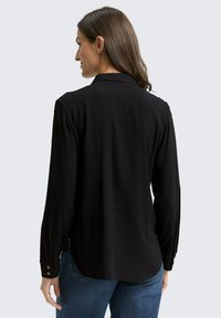 Woman with long brown hair wearing a black long-sleeve blouse and blue jeans, shown from the back against a plain background.