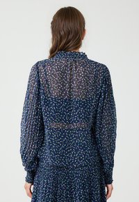Navy doorschijnende blouse met paarse en groene stippen, lange geplooide mouwen, hoge ruchesboord en geweven stof.