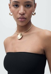Zlatna metalna choker s teksturiranim privjeskom, upotpunjena odgovarajućim zlatnim kružnim naušnicama. Model nosi usku crnu top bez naramenica.
