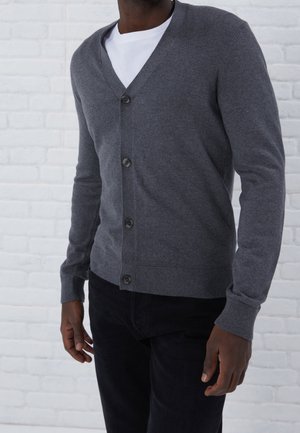 Homme portant un cardigan gris foncé boutonné par-dessus une chemise blanche et un pantalon noir, debout contre un mur en briques blanches.