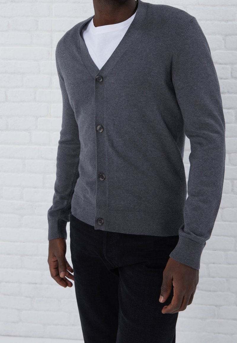 Homme portant un cardigan gris foncé boutonné par-dessus une chemise blanche et un pantalon noir, debout contre un mur en briques blanches.