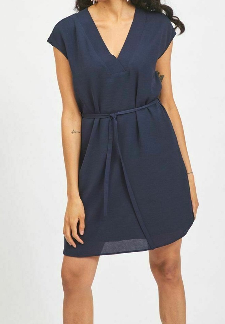 Femme portant une robe bleu marine à col en V avec des manches courtes et une ceinture nouée à la taille, debout devant un fond clair uni.