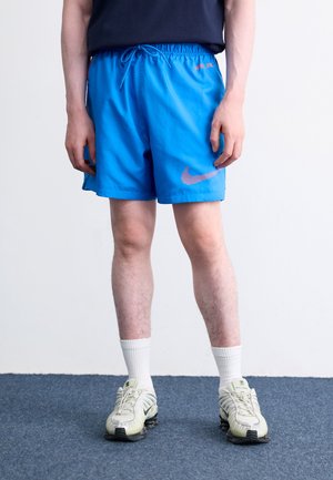 Shorts atletici albaștri cu talie elastică și șnur, având un logo Nike roz. Purtați cu șosete albe și adidași de culoare deschisă.