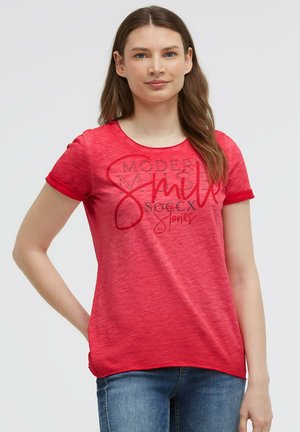 Junge Frau mit langen braunen Haaren trägt ein rotes T-Shirt mit Grafik und blaue Jeans, steht mit einer Hand in der Hüfte vor einem schlichten Hintergrund.