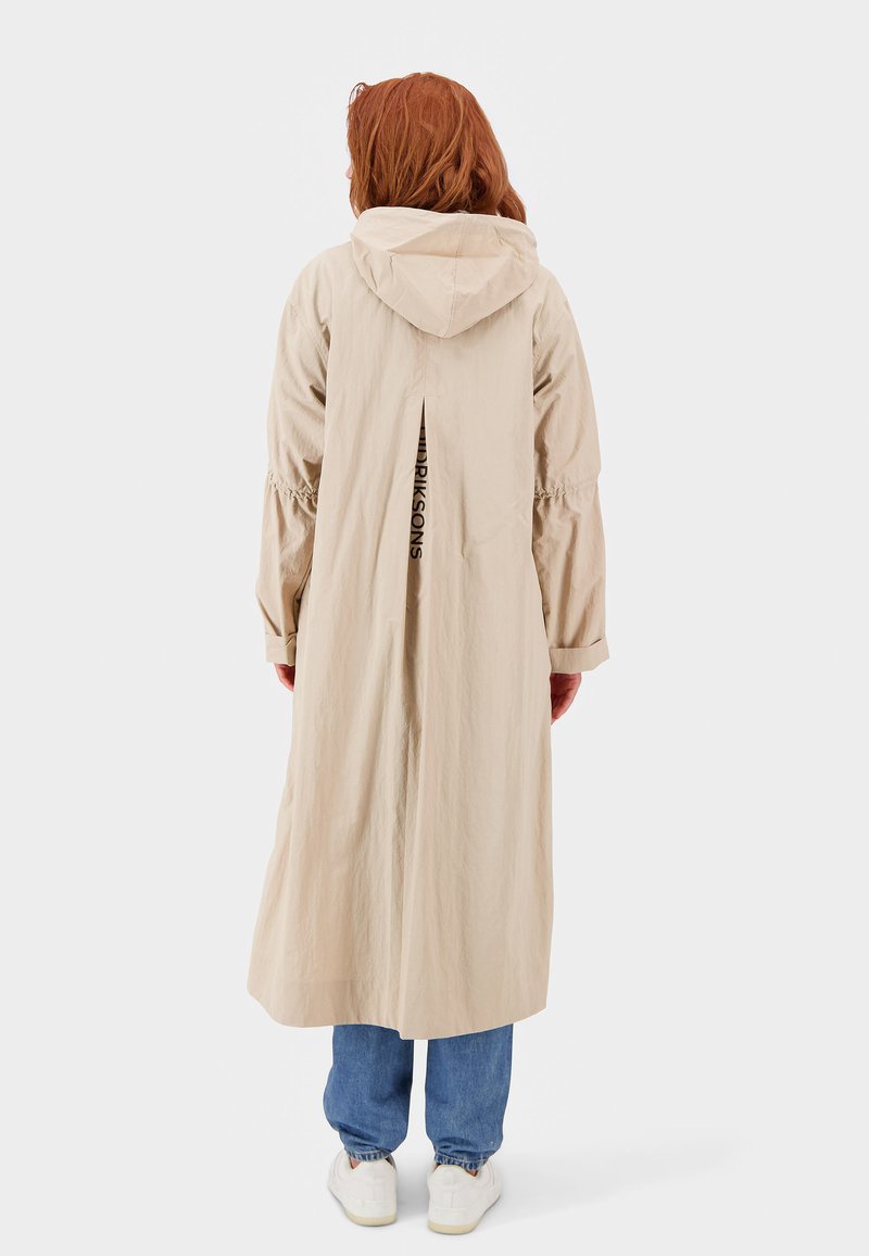 Beige, langer, kapuzen Trenchcoat mit elastischen Bündchen, Rückenbelüftung und Logodetail. Getragen über blauen Jeans und weißen Turnschuhen. Glatte Textur.