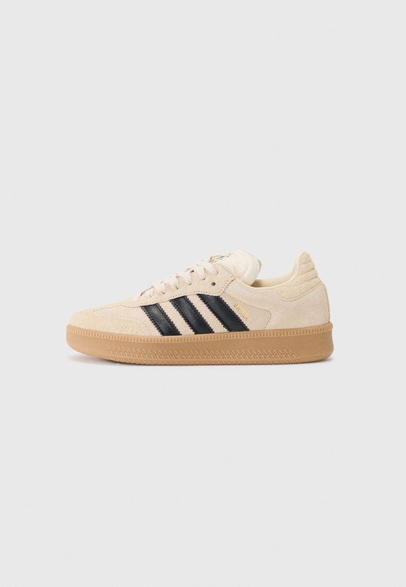 Beige suede sneaker with black stripes, rubber sole, textured accents, and padded collar. Gold logo detail near the heel.