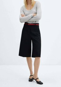 Černé culoty s červeným opaskem, v kombinaci se světle šedým žebrovaným topem s dlouhým rukávem a černými lodičkami, na jednoduchém pozadí.