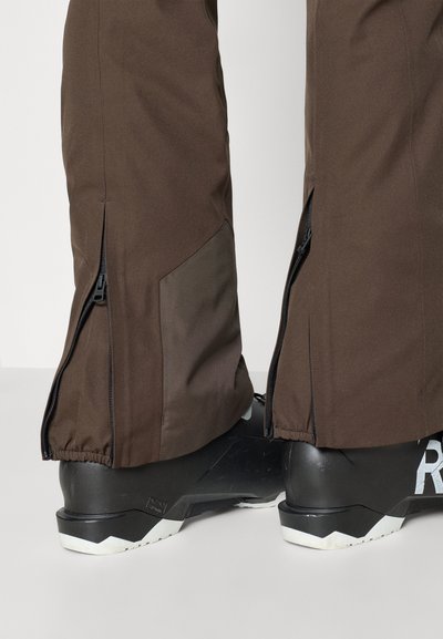 Pruunid veekindlad suusapüksid, millel on lukud pahkluude juures, külgedel tekstureeritud paneel ning neid kannavad mustad suusaätud.