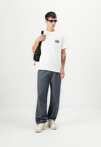 Witte t-shirt met een cirkelvormige graphic, zwarte schoudertas, grijze wijde broek en beige sneakers. Model staat tegen een simpele achtergrond.