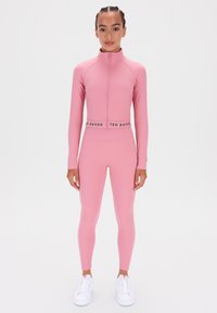 Rózsaszín aktív öltözék szett, amely tartalmaz egy állógalléros zipzáras dzsekit és magas derekú leggingset, rugalmas anyagból készült, sima textúrával és logó részletekkel.