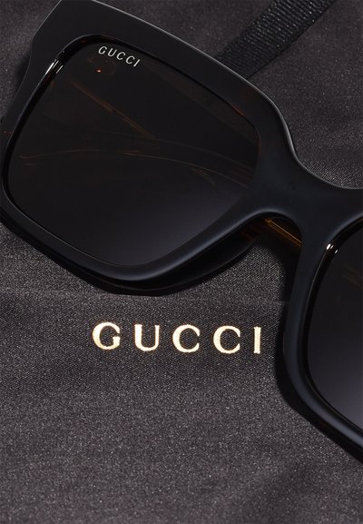 Store svarte solbriller med mørke linser og glatte plastinnramming, plassert på en svart stoff, med den gullfargede "GUCCI"-logoen.