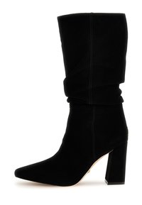 Zwarte, nonchalante enkellaars van zacht suède met een puntige neus en dikke blokhak. Heeft zichtbare sierstiksels en een subtiel gouden accent.