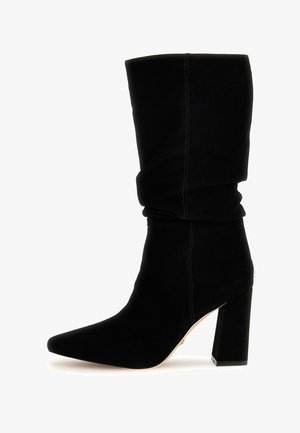 Botín negro y holgado de ante suave con punta puntiaguda y tacón grueso de bloque. Presenta costuras visibles y un sutil detalle dorado.