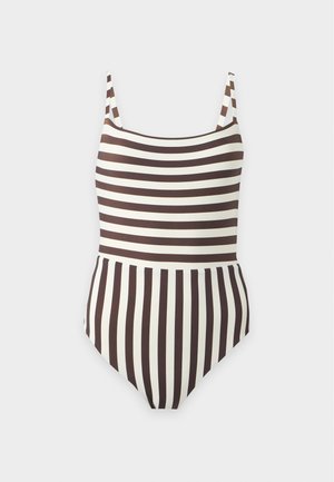 Femme debout pieds nus portant un maillot de bain une pièce rayé noir et blanc avec de fines bretelles sur un fond clair uni.