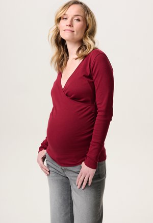 Schwangere Frau mit blonden, welligen Haaren, die ein langärmliges rotes Oberteil und graue Jeans trägt, vor einem schlichten hellen Hintergrund steht.