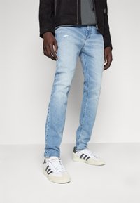 Jeans de denim azul claro con un corte ajustado, que presentan un detalle desgastado en la parte superior del muslo, combinados con zapatillas blancas y grises.