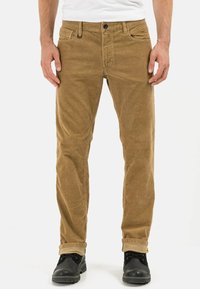 Pantalon en velours côtelé beige à coupe droite, avec poches avant, fermeture à boutons et revers retroussés. Porté avec des chaussures noires.