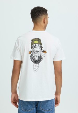 Homme portant un t-shirt blanc avec un motif d'une girafe portant un bonnet olive vert étiqueté "COOL" et une grosse écharpe noire.